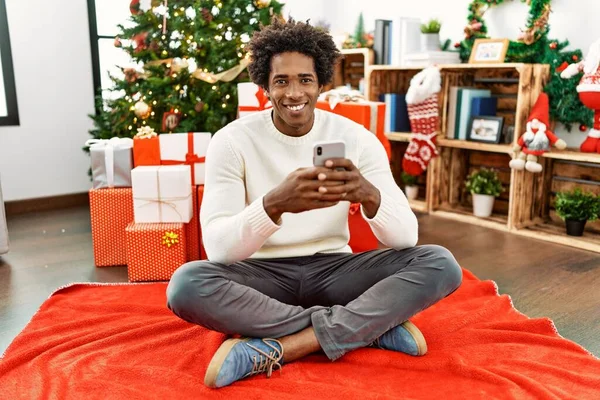 Jovem Afro Americano Usando Smartphone Sentado Chão Pela Árvore Natal — Fotografia de Stock