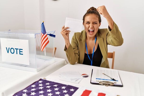 Красивая латиноамериканка, держащая избирательный конверт в урне для голосования, раздраженная и разочарованная, кричащая от гнева, кричащая безумно от злости и поднятой руки 