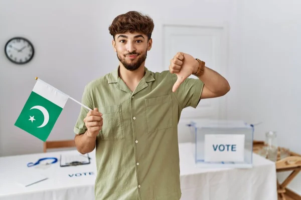 Young arab man at political campaign election holding pakistan flag with angry face, negative sign showing dislike with thumbs down, rejection concept