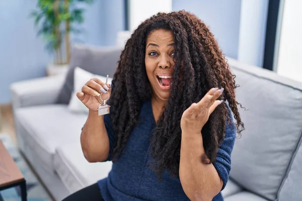 Plus size hispanic woman holding keys of new home celebrating achievement with happy smile and winner expression with raised hand