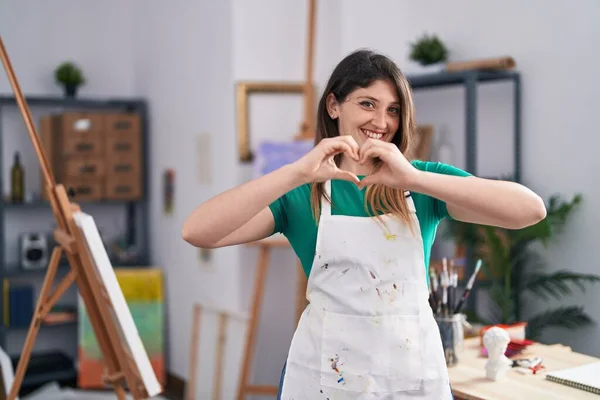 Young brunette woman at art studio smiling in love doing heart symbol shape with hands. romantic concept.