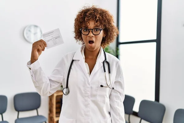 Young african american doctor woman holding covid record card scared and amazed with open mouth for surprise, disbelief face