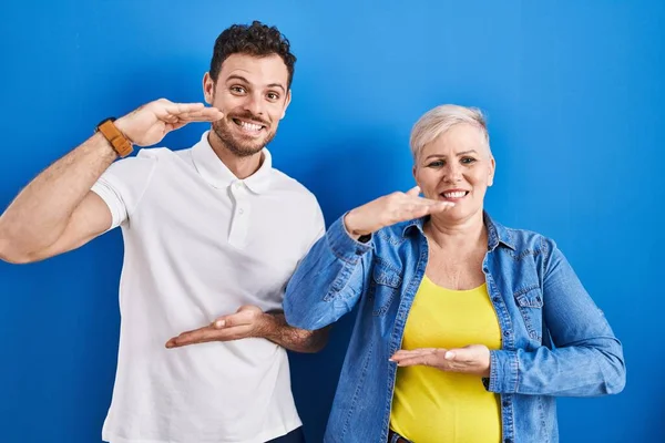 若いブラジル人の母親と息子の手で青の背景ジェスチャーの上に立って大小のサインを示す 指標のシンボル カメラを見て笑ってる 概念を測定し — ストック写真