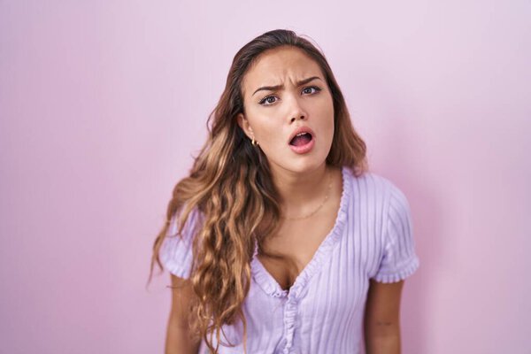 Young hispanic woman standing over pink background in shock face, looking skeptical and sarcastic, surprised with open mouth 
