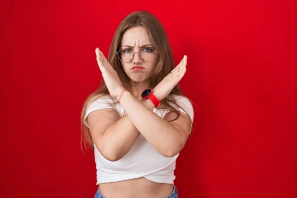 年轻的高加索女人站在红底排斥表情旁边 交叉着双臂做着消极的手势 满脸怒容 — 图库照片