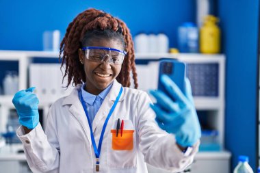 African woman working at scientist laboratory doing video call screaming proud, celebrating victory and success very excited with raised arm 