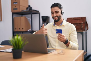 Çağrı merkezi kulaklığı takan İspanyol genç adam online alışveriş yapıyor mutlu ve olumlu gülümsüyor, başparmağıyla mükemmel ve onay işareti yapıyor. 