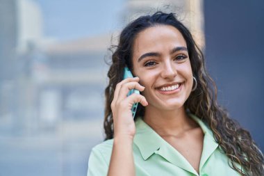 Genç, güzel, İspanyol bir kadın gülümsüyor ve caddede akıllı telefondan konuşuyor.