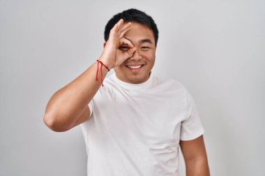 Young chinese man standing over white background doing ok gesture with hand smiling, eye looking through fingers with happy face. 