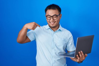 Chinese young man using computer laptop looking confident with smile on face, pointing oneself with fingers proud and happy. 