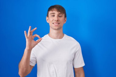Caucasian blond man standing over blue background smiling positive doing ok sign with hand and fingers. successful expression. 