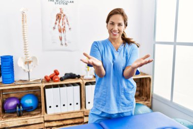 Middle age hispanic physiotherapist woman working at pain recovery clinic smiling cheerful offering hands giving assistance and acceptance. 