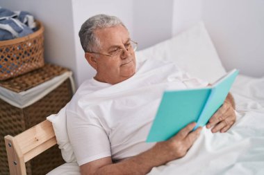 Orta yaşlı, kır saçlı, kitap okuyan, yatak odasında oturan bir adam.