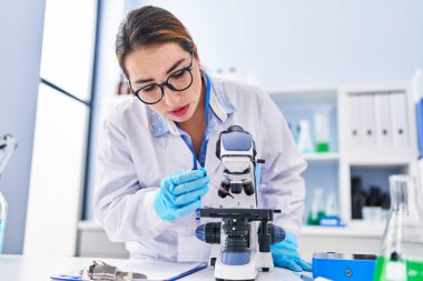 Young beautiful hispanic woman scientist using microscope pouring blood on sample at laboratory