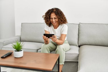 Orta yaşlı İspanyol kadın evde akıllı telefon kullanarak gülümsüyor.