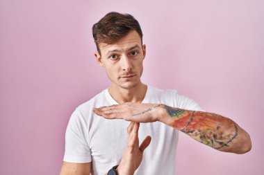 Caucasian man standing over pink background doing time out gesture with hands, frustrated and serious face 