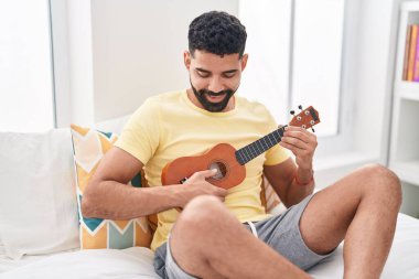Young arab man playing ukulele sitting on bed at bedroom