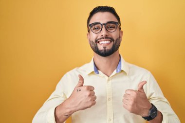 İspanyol genç adam iş kıyafetleri ve gözlük takıyor. Başarı işareti ile el hareketi yapıyor. Gülümsüyor ve mutlu. Neşeli ifade ve kazanan jest. 