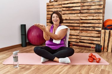 Middle age hispanic woman sitting on training mat at the gym pointing aside with hands open palms showing copy space, presenting advertisement smiling excited happy 