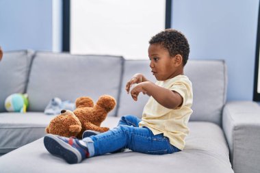 Afrikalı Amerikalı çocuk evde kanepede oyuncak ayıyla oynuyor.