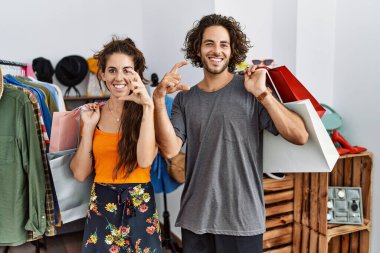 Satış mağazasında alışveriş torbaları tutan genç İspanyol bir çift gülümsüyor ve el hareketleriyle el hareketleriyle küçük boy işareti yapıyor ve kameraya bakıyor. ölçüm kavramı. 