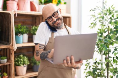 Genç, kel bir çiçekçi çiçekçide bilgisayarla akıllı telefondan konuşuyor.