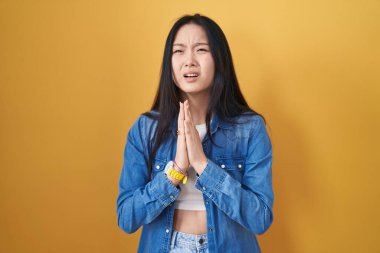 Sarı arka planda duran Asyalı genç bir kadın elleri bağlı yalvarıyor ve dua ediyor. Yüzünde umut ifadesi var. Çok duygusal ve endişeli. Yalvarıyor. 