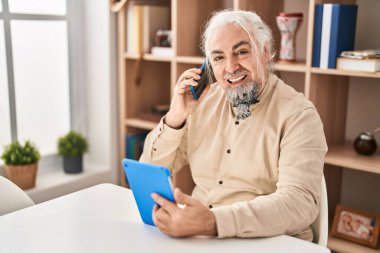 Middle age grey-haired man talking on smartphone using touchpad at home