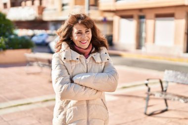 Orta yaşlı kadın gülümsüyor. Kollarını kavuşturup caddede duruyor.