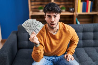 Sakallı İspanyol bir adam elinde 100 dolarlık banknotlar tutuyor. Ağzını açıp şaşırmış ve korkmuş. 