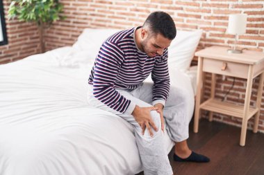 Young hispanic man suffering for knee injury sitting on bed at bedroom