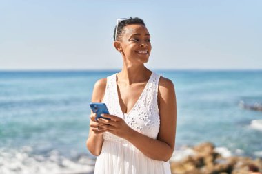 Afrika kökenli Amerikalı bir kadın deniz kenarında akıllı telefon kullanarak gülümsüyor.