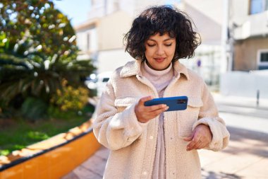 Genç, güzel, İspanyol bir kadın, gülümsüyor. Parkta akıllı telefondan video izliyor.