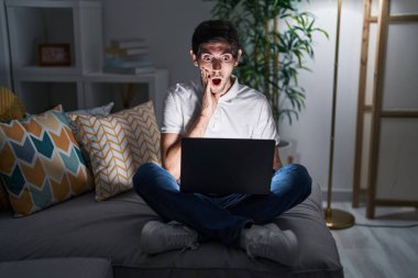 Young hispanic man using laptop at home at night afraid and shocked, surprise and amazed expression with hands on face 