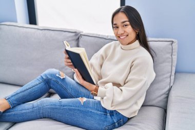 Evdeki kanepede oturmuş kitap okuyan genç İspanyol kadın.