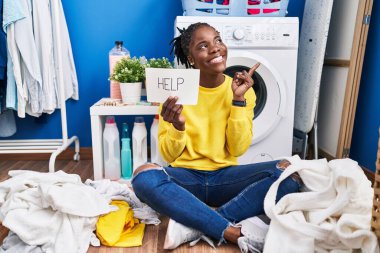 Beautiful black woman doing laundry asking for help smiling happy pointing with hand and finger to the side 