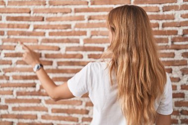 Young caucasian woman standing over bricks wall posing backwards pointing ahead with finger hand 