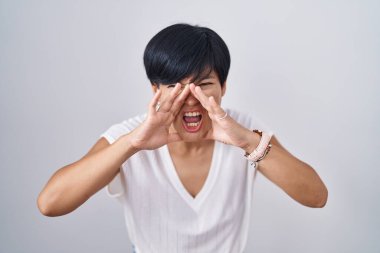 Young asian woman with short hair standing over isolated background shouting angry out loud with hands over mouth 