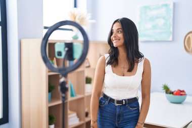 Brunette woman recording vlog tutorial with smartphone at home looking to side, relax profile pose with natural face and confident smile. 
