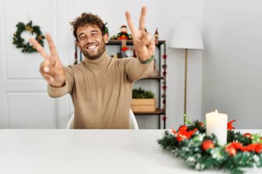 Noel süslemesi masasında oturan sakallı yakışıklı genç adam iki elini de zafer işareti yaparken göstererek gülümsüyor. İki numara.. 