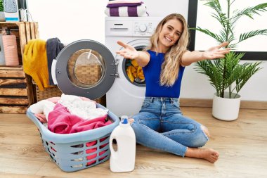 Çamaşır sepetinin yanında oturan genç ve güzel bir kadın kucaklanmak için kollarını açıp kameraya bakıp gülümsüyor. Mutluluğu kucaklayan neşeli bir ifade.. 