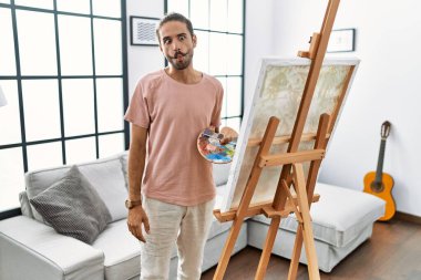 Young hispanic man with beard painting on canvas at home making fish face with lips, crazy and comical gesture. funny expression. 