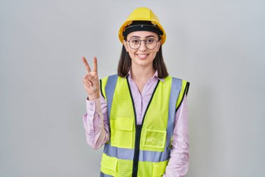 Hispanic girl wearing builder uniform and hardhat showing and pointing up with fingers number two while smiling confident and happy. 