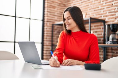 Young beautiful hispanic woman using laptop writing on document at home