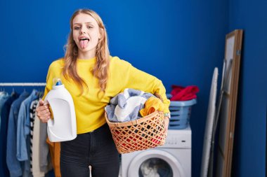 Young caucasian woman holding laundry basket and detergent bottle sticking tongue out happy with funny expression. 