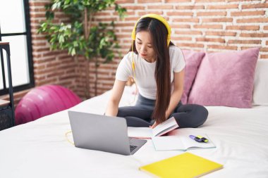 Young chinese woman student writing on book studying on bed at bedroom