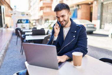 Genç İspanyol adam akıllı telefondan konuşuyor. Kahve dükkanının terasında dizüstü bilgisayar kullanıyor.