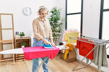 Young caucasian woman listening to music ironing clothes at laundry room