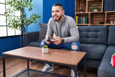 Young latin man playing video game sitting on sofa at home