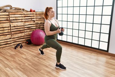 Genç kızıl saçlı kadın spor merkezinde kettlebell ile antrenman yapıyor.